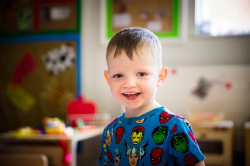 A child is smiling at the camera. He has short blond hair and is wearing a blue t-shirt with superhero faces on it.