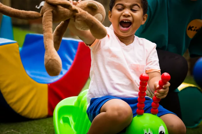 Joseph is sitting on a caterpillar rider. He is holding a teddy in the air while shouting/laughing.