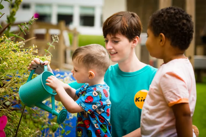 A child is watering a bush with help from a staff member. There is another child watching them on the right.
