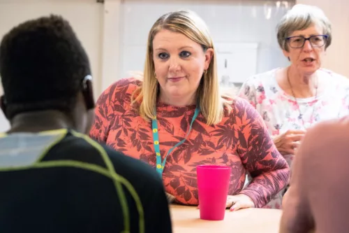 A staff member is looking at a young person whose back is facing the camera. They are both sitting around a table. There is another member of staff in the background.