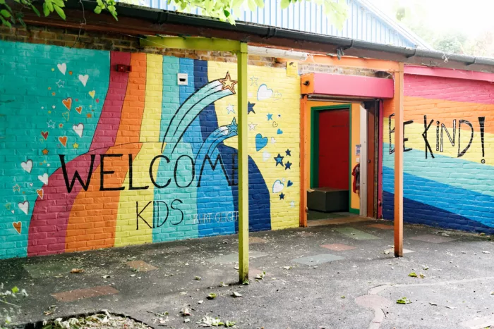 The front building of the Hackney Playground is painted magenta, orange, yellow, green, light blue and dark blue. On the wall there is writing which reads, 'Welcome Kids - be kind'