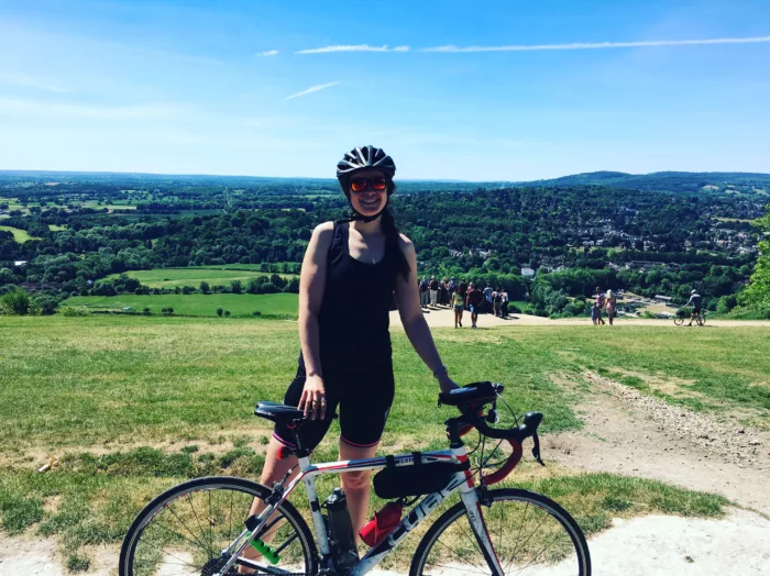 Josie is in her sports gear, holding onto her bike. She is in a green area with a forest in the background. There are other people grouped together and holding bikes.