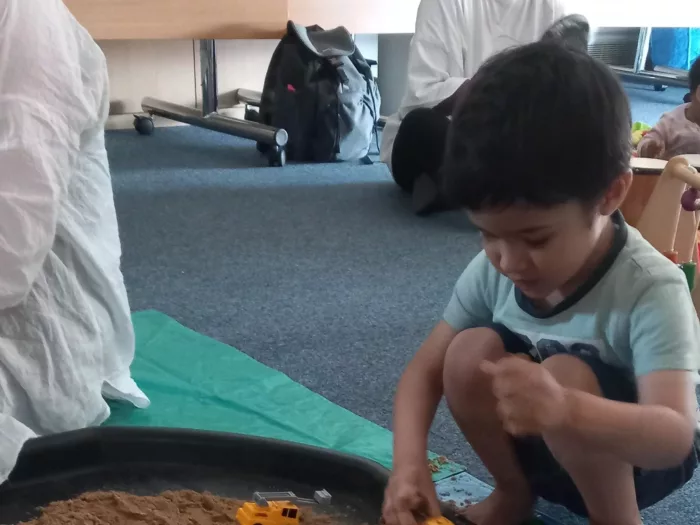 Hamza is crouched on the floor, playing with trucks in a sand tray.