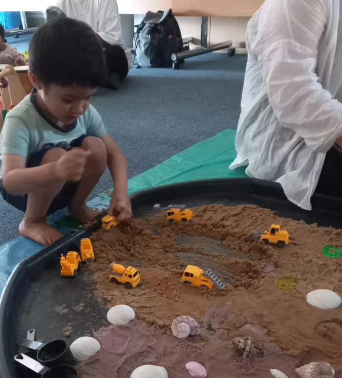 Hamza is crouched on the floor, playing with trucks in a sand tray.