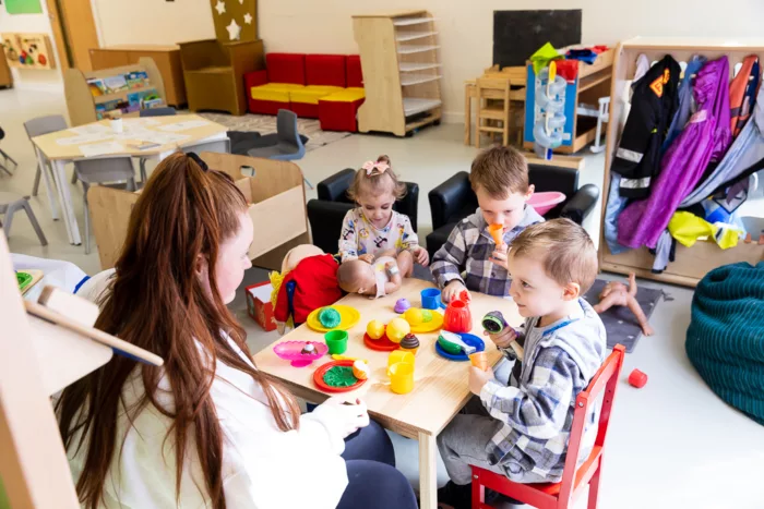 Three children are sitting around a kids table with a staff member. They are pretending to have lunch with toys.