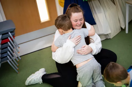 A child is photographed from behind, hugging a staff member who is kneeling on the floor.