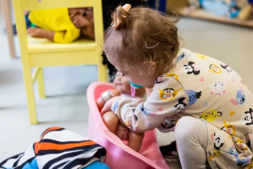 A girl is photographed from the side. She is putting a fake baby into a baby bathtub.