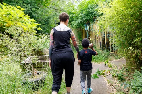 A staff member is walking with a child in a green area. They are photographed from behind.