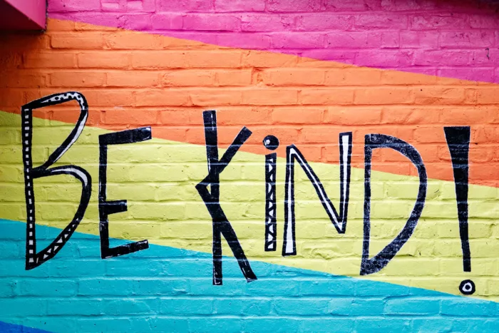 A wall painted magenta, orange, yellow and blue. There is text painted on top, which reads ' Be kind'.