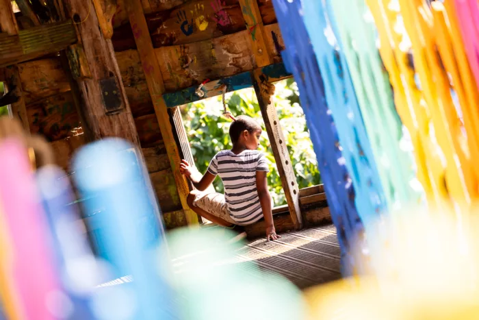 A boy is inside a playground playhouse. He is sitting by the window, his back to the camera.
