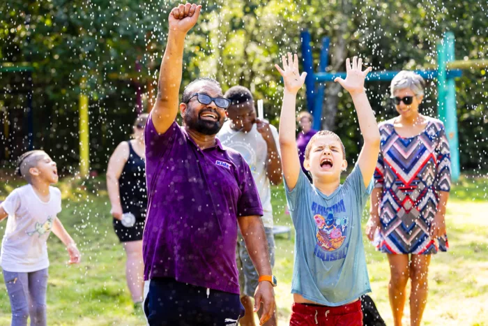 A staff member and a child are standing outside with their arms up in the air. There are water drops falling on them.