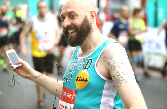 Terry is at the London Marathon, holding onto his phone with earphones in one ear. He is wearing a blue vest with the old Kids logo on it.