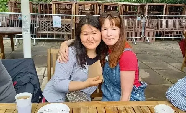 One woman is leaning on another woman, who has an arm wrapped around her. They are sitting at an outdoor table.