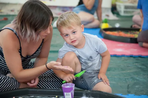 A boy is kneeling in front of a sensory play tray. There is a woman next to him