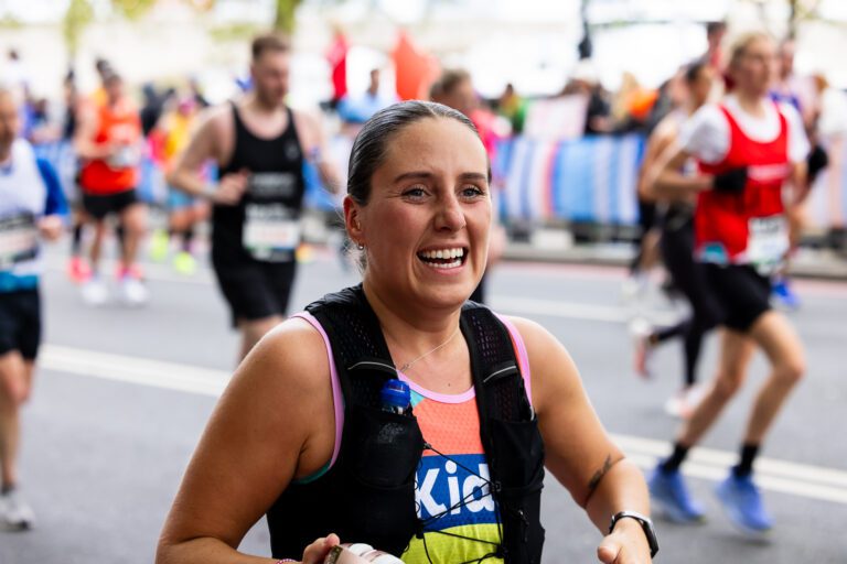 A Woman is running in a Kids vest. She is running with both hands up in the air