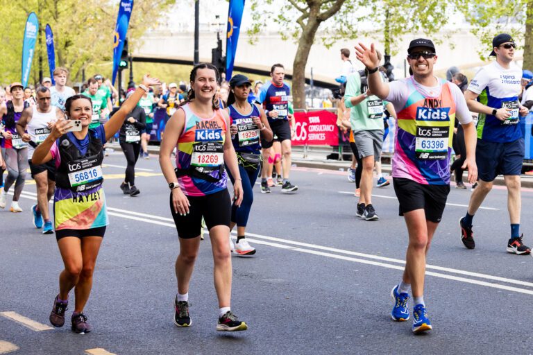 Three people are walking during the London Marathon. They are all wearing Kids vests