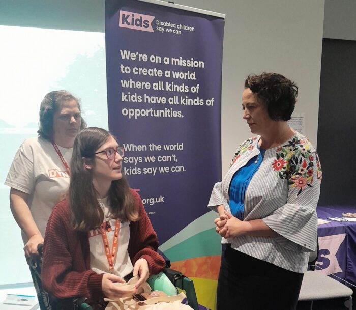 3 woman talking in front of a Kids branded banner. One woman is a wheelchair user.